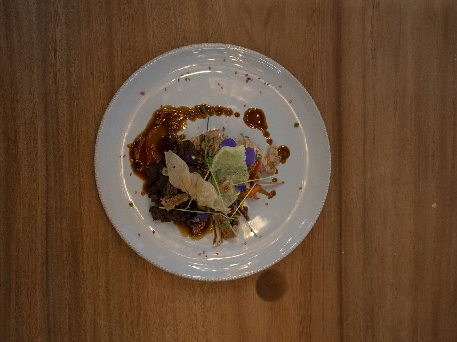 Een bord met vlees, groenten en een sausstreep op een houten tafel, van bovenaf gezien